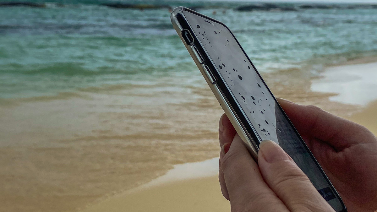 A wet smartphone with water droplets on the screen, held in someone's hands on a beach.